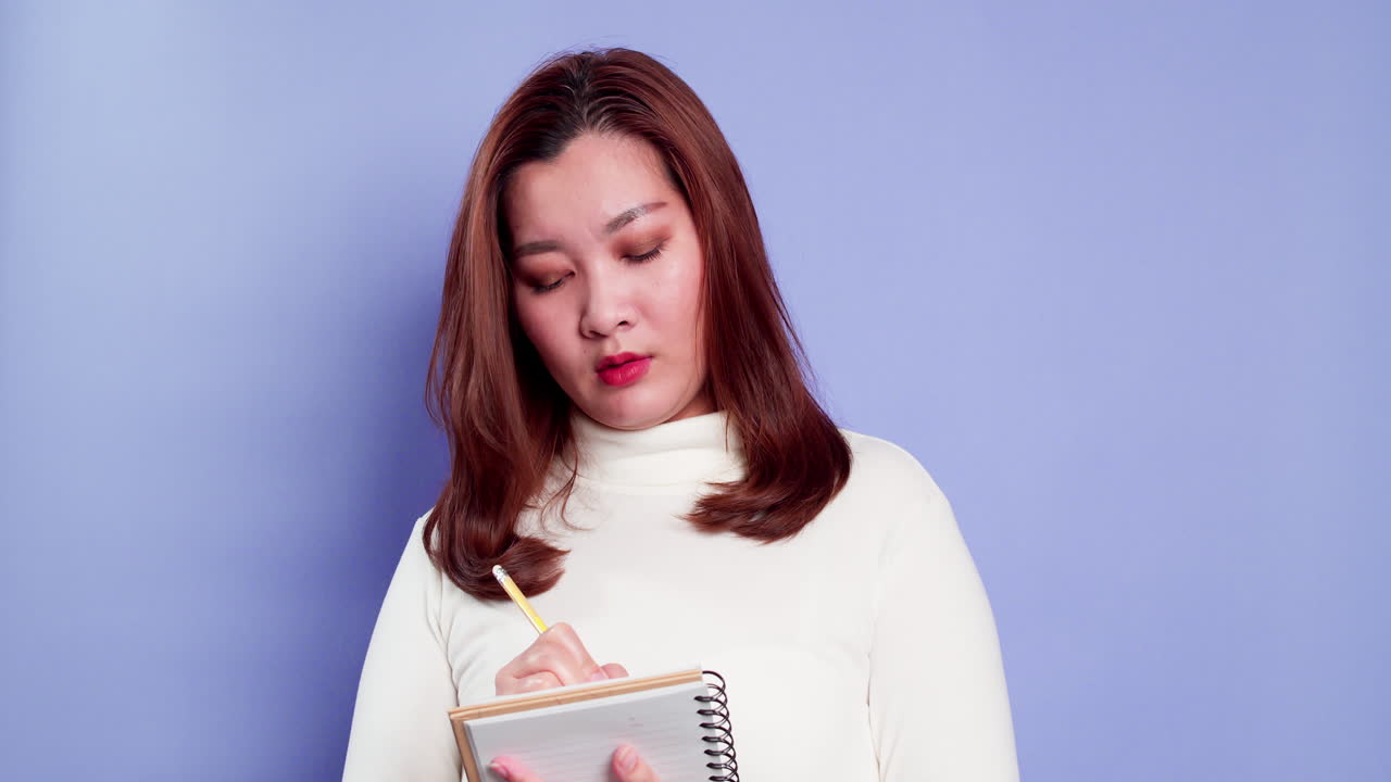 Happy beautiful Asian girl making notes in a paper notebook, writing creative ideas and to-do list indoor studio shot isolated on purple background