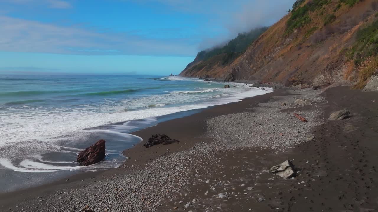 Waves crashing surf black sand rugged coastline sunny morning blue sky mist fog layer USAL Beach Campground Lost Coast Trail California aerial drone PNW boulders rocks mountainside upwards motion