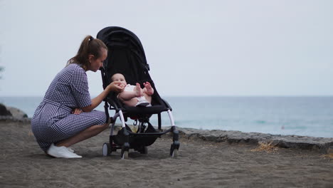 Frente-A-La-Serena-Vista-Del-Océano,-Una-Joven-Madre-Interactúa-Con-Su-Bebé-En-El-Cochecito,-Encarnando-El-Cuidado-Y-El-Afecto-Dentro-De-Esta-Dulce-Y-Joven-Familia.