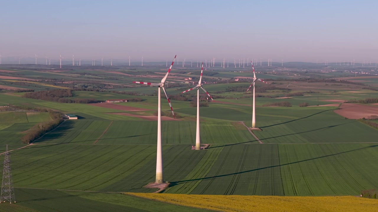 Luchtbeelden van een windmolenpark in een agrarisch landschap. Meerdere witte windmolens met rode markeringen in groene en bruine velden. Installatie voor hernieuwbare energie op het platteland.