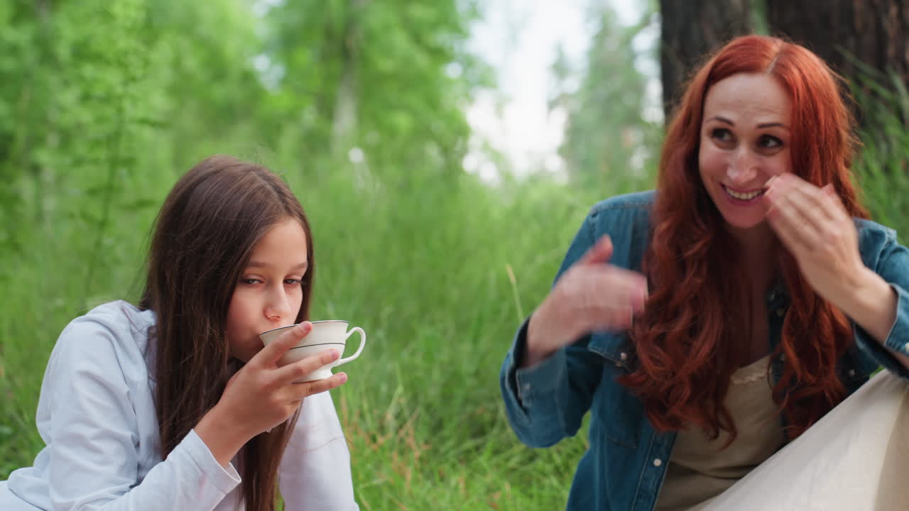 엄마와 딸이 피크닉 담요에 앉아 즐겁게 이야기를 나누고 있어요. 딸은 컵으로 차를 마시고 엄마는 재미있는 표정을 짓고 있으며, 주변은 푸른 잔디와 따뜻한 자연광으로 둘러싸여 있어요.