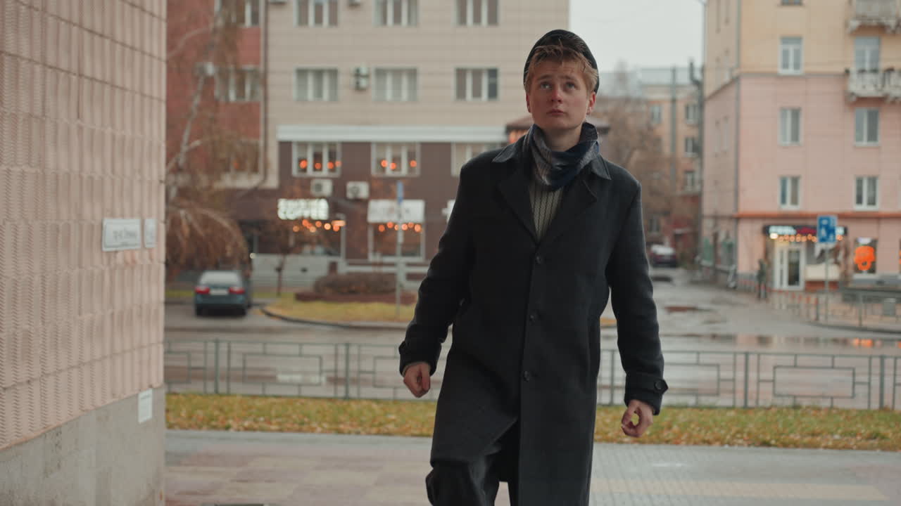 Giovane creatore caucasico in piedi sotto la pioggia su gradini urbani, che si avvicina a un ingresso, cappotto e sciarpa bagnati, basco inclinato, sguardo contemplativo verso il cielo, color grading cinematografico, atmosfera autunnale sommessa.