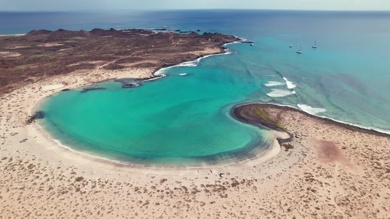 Aerial 4k drone shot of Playa De La Concha De Lobos on Lobos Island, showcasing volcanic landscapes, a crescent-shaped beach cove, and vivid turquoise waters under a bright, clear sky.
