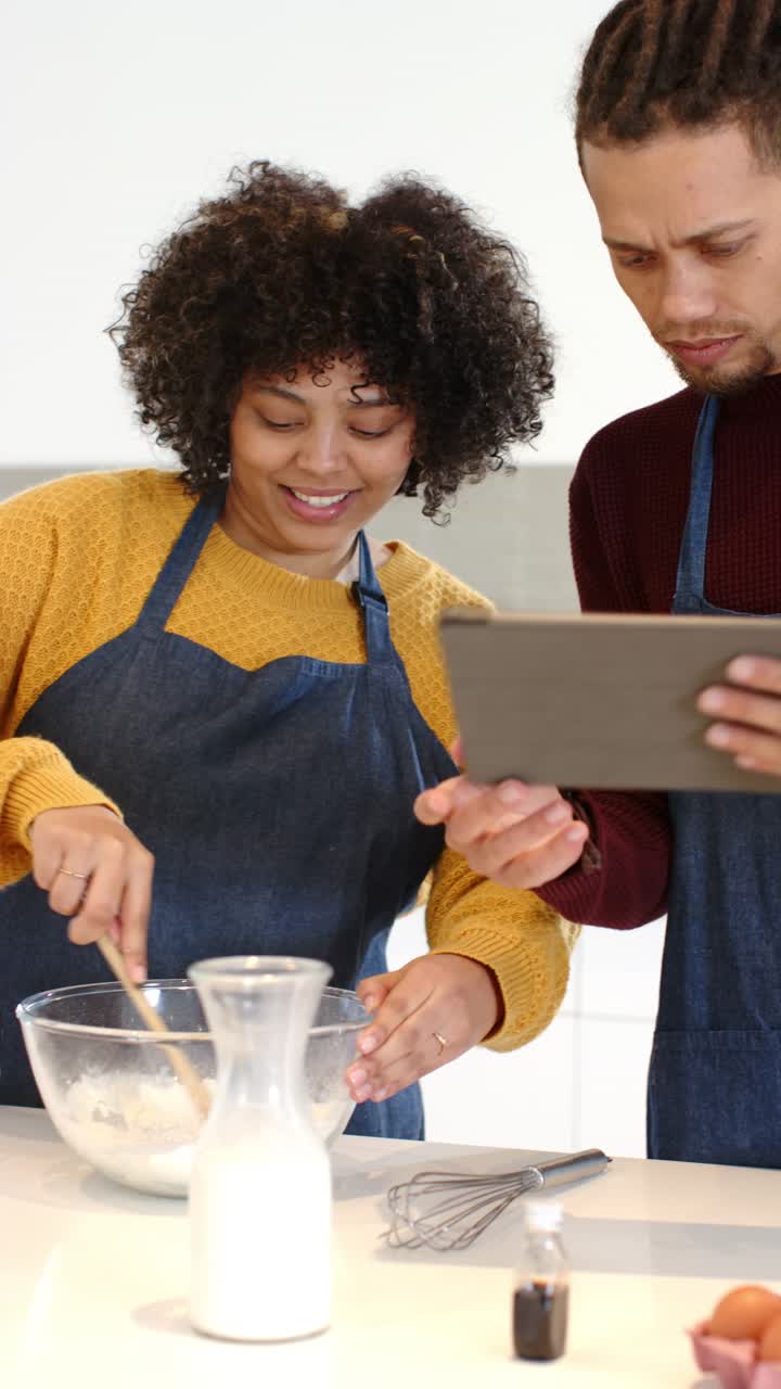 Vertical video: Diverse couple stirring batter with wooden spoon in kitchen following tablet steps