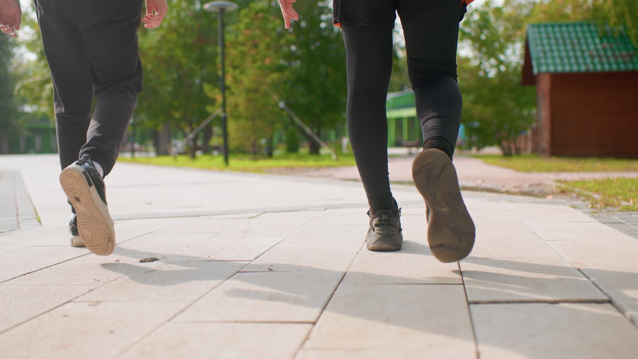 low angle white men walking forward, tight focus on legs and feet in sneakers with one using skateboard, tiled pavement and sunlit park setting, rhythmic steps and momentum, roles suggested courier