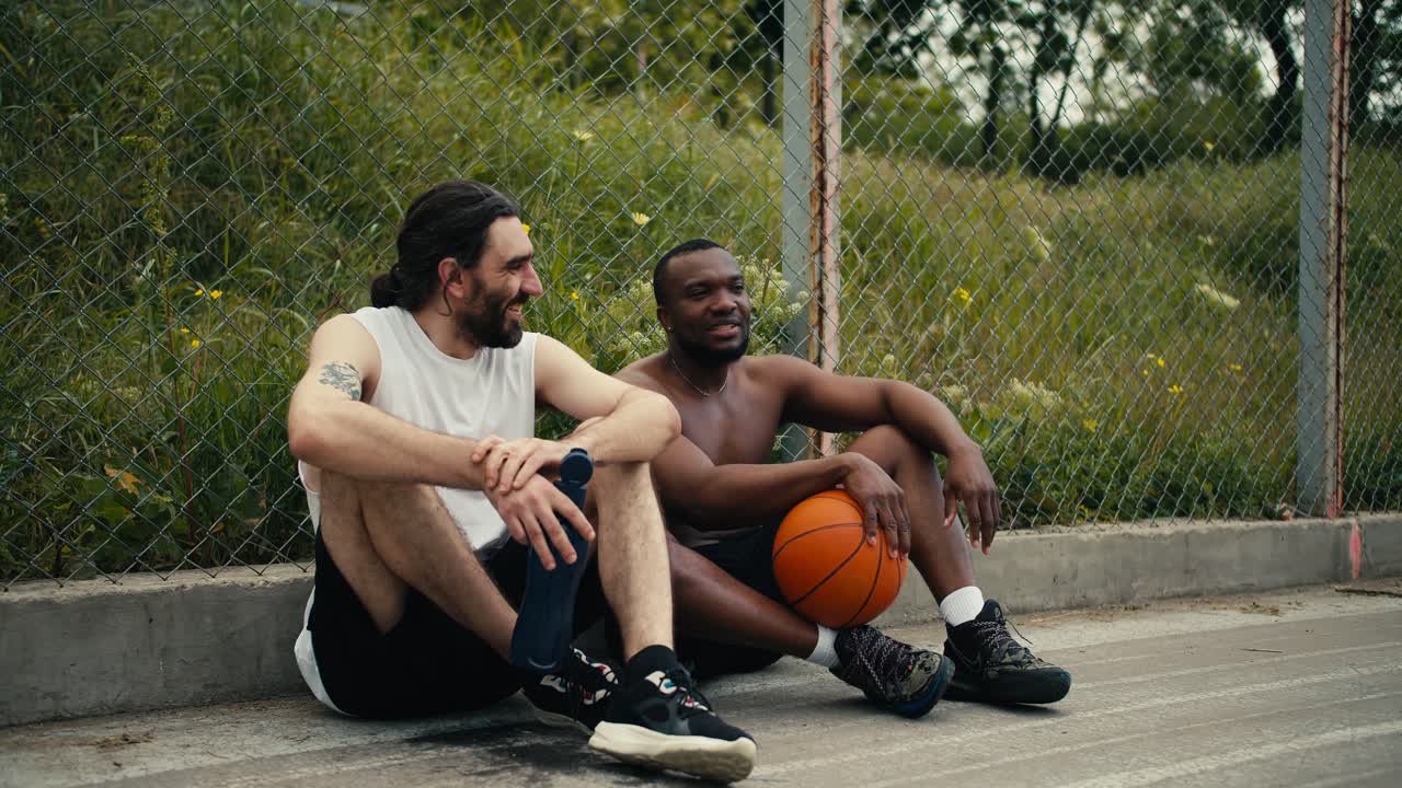 Two men are sitting on the floor of the basketball court and chatting about the ongoing match, they are happy that they can attend the game