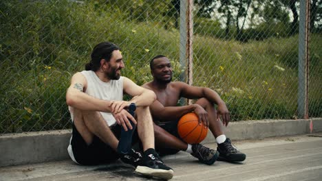 Zwei-Männer-Sitzen-Auf-Dem-Boden-Des-Basketballplatzes-Und-Unterhalten-Sich-über-Das-Laufende-Spiel,-Sie-Freuen-Sich,-Dass-Sie-Dem-Spiel-Beiwohnen-Können