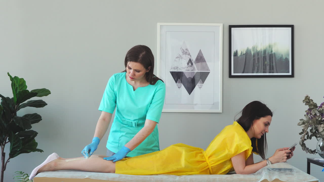 la chica habla por teléfono y sonríe durante la depilación de las piernas en el salón de belleza. depilación de las piernas con azúcar. maestro de depilación