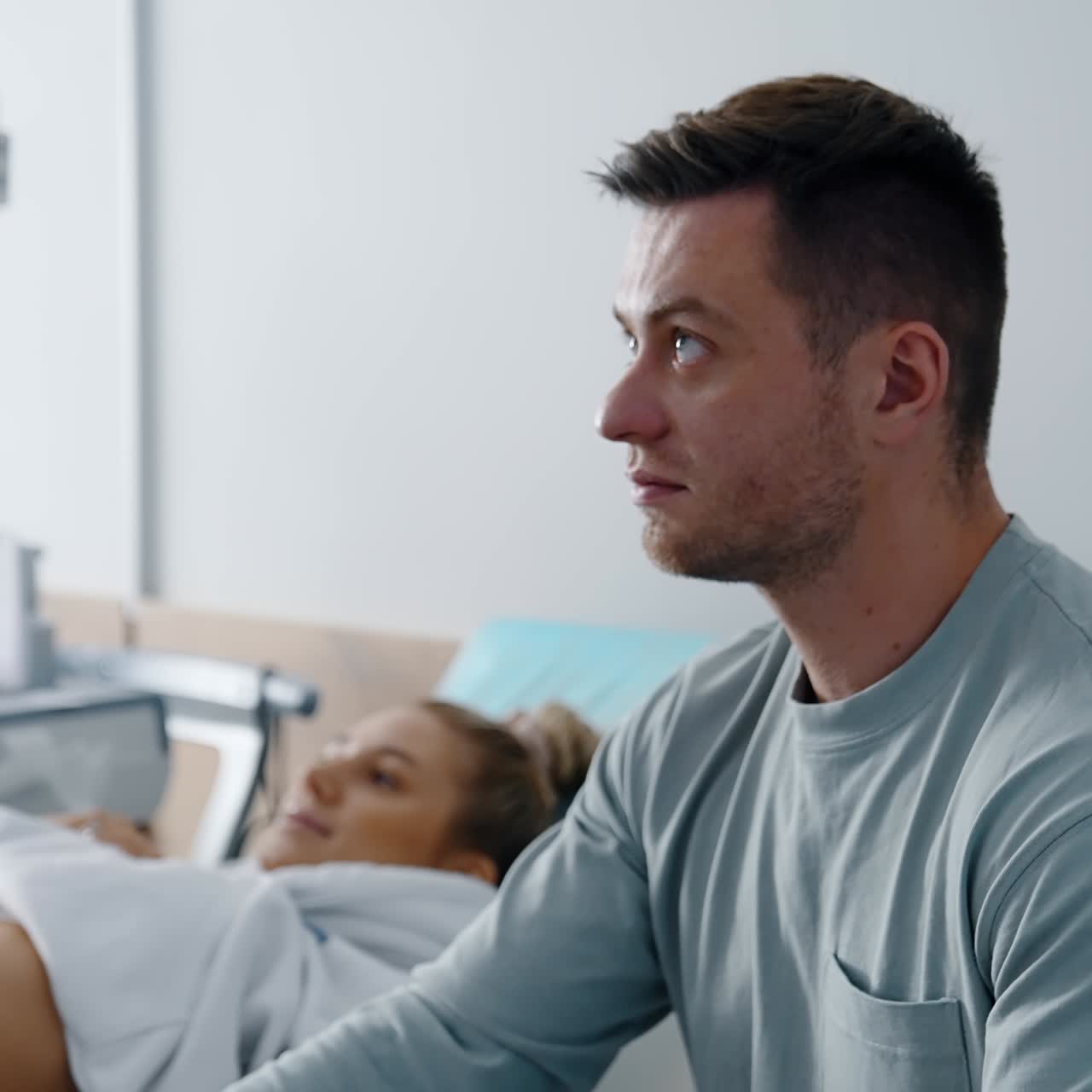 Uomo barbuto concentrato che guarda lo schermo che mostra il suo futuro bambino. Donna bionda sdraiata sul divano sullo sfondo, in secondo piano