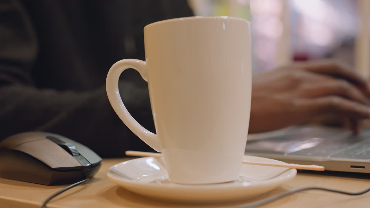 Tazza bianca in primo piano sulla scrivania, laptop sfocato e mani che digitano sullo sfondo, luce interna calda, piattino e mouse visibili, scrittore freelance al laptop che crea un'atmosfera calma e produttiva.