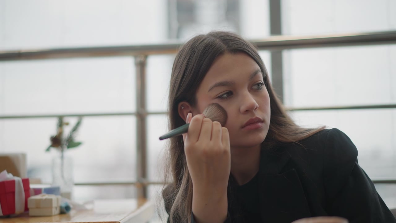 Young woman seated at table applies face powder with makeup brush, gently turning head and raising hand to check skin result beside window in soft natural light, with woman, makeup, brush, powder