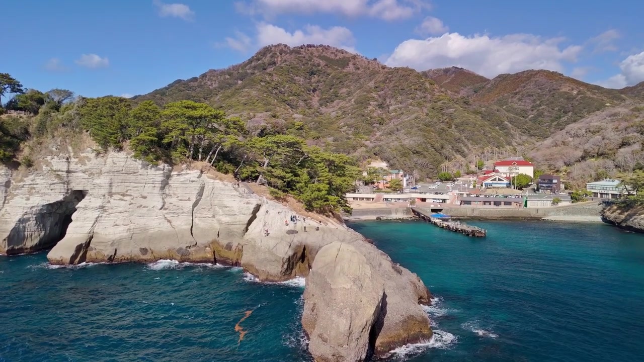 Sweeping aerial view of a dramatic rocky cape, sea cave entrances, and a quiet harbor village under a clear blue sky on the Izu Peninsula.