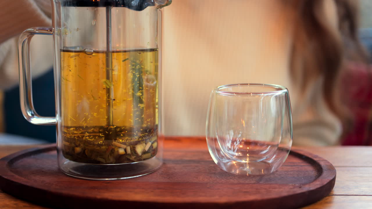Close up of a woman pressing down the tea leaves with the help of a French press near a cup of tea at a restaurant