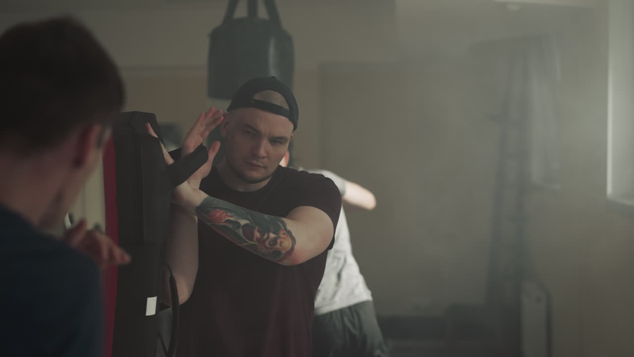 팔에 문신을 한 전문 트레이너는 학생이 어두운 체육관에서 키크박싱을 연습하기 위해 권투 장비를 들고 있습니다. 코치와 함께 공격적인 종류의 스포츠를 연습하기위한 싸움 클럽