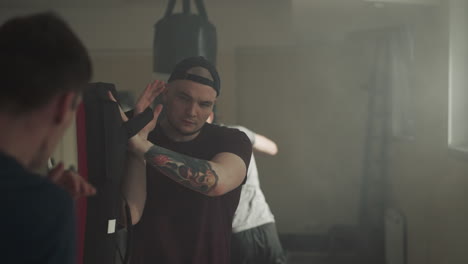 professional trainer with tattoo on arm holds boxing equipment for student to practice kickboxing in dark gym. fight club for practicing aggressive types of sports with coach