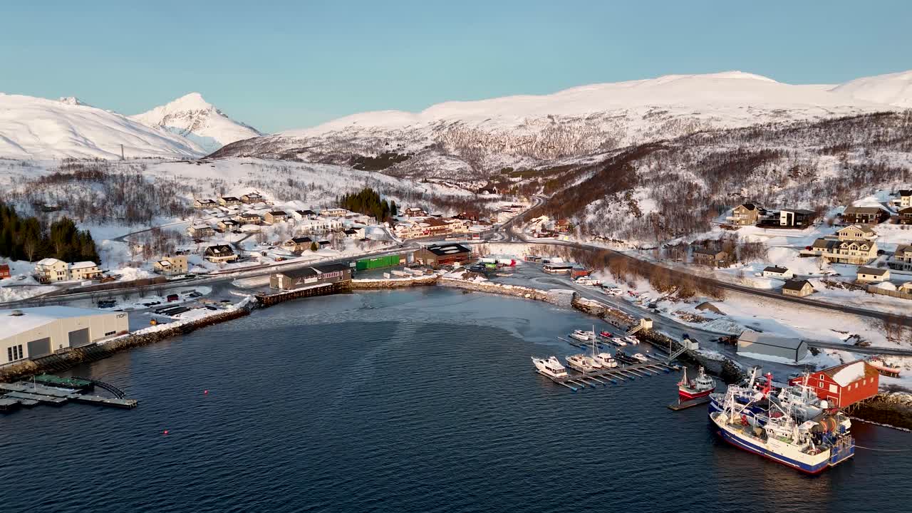 Drohnen-Panoramaansicht über einem Küstenhafen und Wohngebiet nahe Tromsø, Norwegen. Fischerboote, Gebäude am Wasser und schneebedeckte arktische Berge im winterlichen Tageslicht.