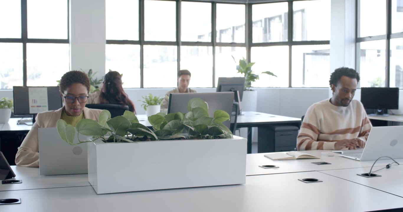 Un team eterogeneo in ufficio; una panoramica della telecamera rivela una donna in blazer che digita su un laptop accanto a una fioriera, intenta a lavorare.