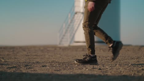 Worker-in-boots-walks-past-a-wind-turbine-on-a-clear-day,-with-a-focus-on-the-steps-and-turbine-base