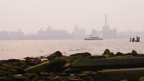 View-of-Manhattan-covered-in-smoke-from-wildfires-seen-from-beach-on-the-east-river-with-waves-crashing-on-mossy-rocks-in-the-foreground-and-a-ferry-crossing-frame