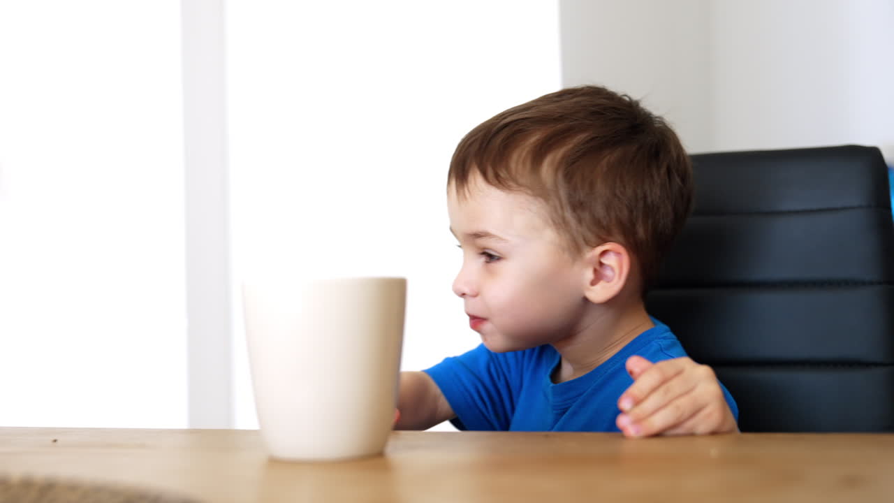 Smiling child enjoying a drink at home. A young boy in a blue shirt sits at a wooden table, happily tasting his drink with an expression of delight