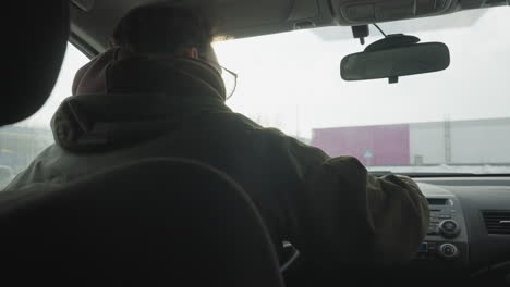 rear view of man silhouetted by sunlight cleaning car dashboard controls with blue cloth, distant pink and white building and sign seen through snowy windshield in urban parking lot scene