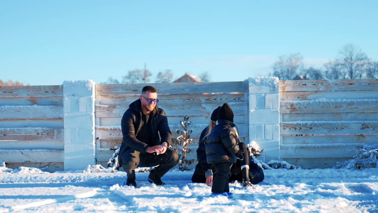 Fashionable Caucasian family outdoors in winter. Father makes a snowball and playfully to a kid.