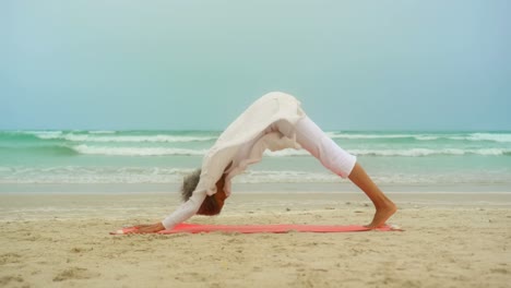 Side-view-of-active-senior-African-American-woman-doing-yoga-on-exercise-mat-at-the-beach-4k