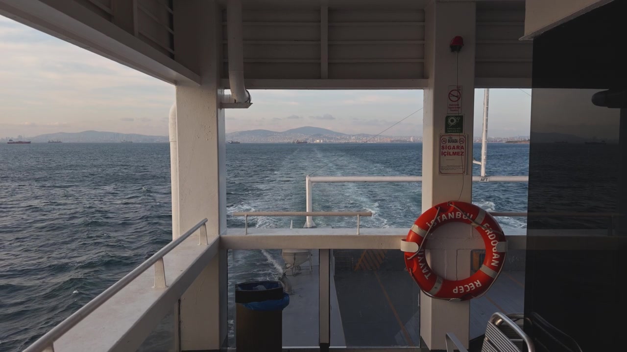 Blick vom Deck einer fahrenden Fähre mit einem orangefarbenen Rettungsring auf einem Pfeiler und dem offenen Meer sowie dem sichtbaren Kielwasser im Hintergrund bei Tageslicht, Szene einer Seereise und Überfahrt in der Türkei.