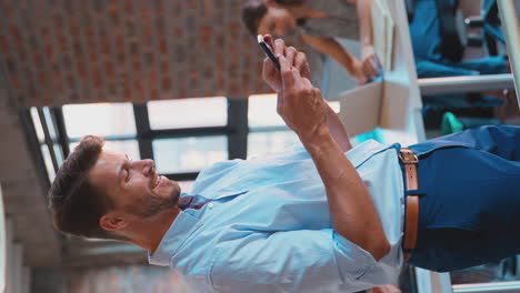 Vertikales-Video-Eines-Lächelnden-Geschäftsmannes,-Der-In-Einem-Geschäftigen-Büro-Steht-Und-Nachrichten-Auf-Dem-Mobiltelefon-Sendet