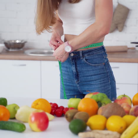 dieting concept. young woman measures her belly in the kitchen. organic fruit and vegetables on the table. model tries to lose her weight. healthy diet.