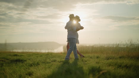 dos parejas bailan felices en una colina cubierta de hierba iluminada por el sol al lado de un lago pacífico. el hombre, vestido con una camisa blanca, sombrero y vaqueros, comparte un momento con su esposa, que lleva un vestido blanco y un sombrero negro