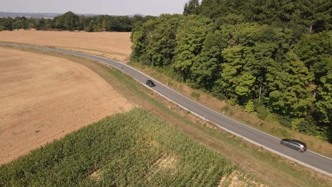 Dos-Coches-Circulando-En-Diferentes-Direcciones-Por-Una-Carretera-Rural-Curva-Entre-Campos-De-Maíz-Y-Bosques-Frondosos