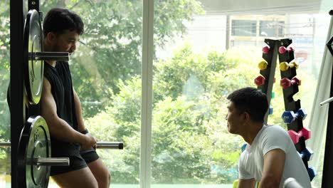 two men engage in conversation during a workout in a well-lit bangkok gym, surrounded by exercise equipment
