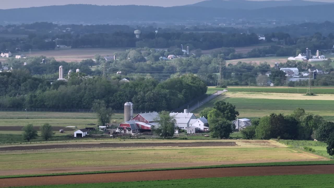 Aerial zoom shot of industrial farmstead in peaceful american suburb area in spring. Wide shot. Forest trees and colored farm fields in rural district of town.