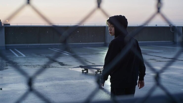 Teenager i hættetrøje kigger mod kameraet, bevæger sig og rækker ud efter et skateboard på et tag