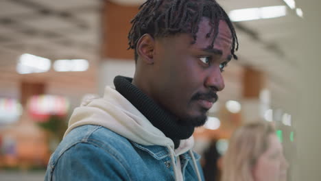 close-up of young man with short hair and braided locks, dressed in denim jacket and white hoodie, staring thoughtfully with a focused expression in indoor public space