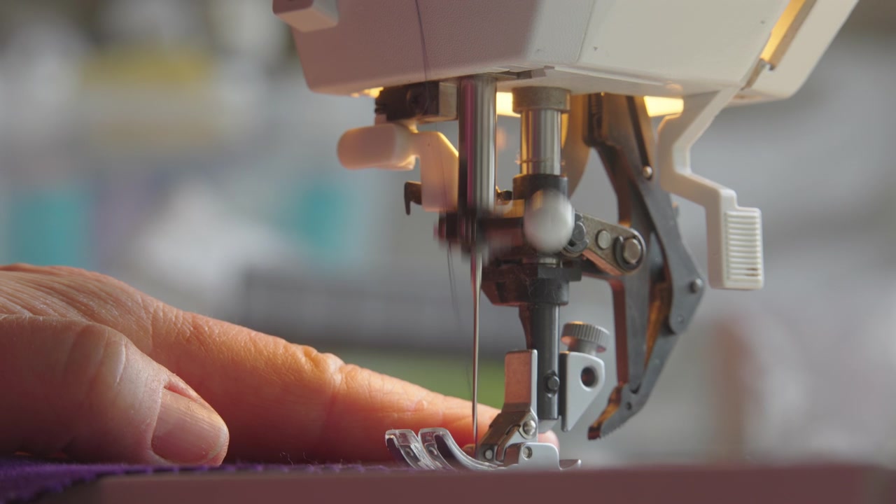Mature hands guide fabric at sewing machine as needle moves up and down during garment construction, close up
