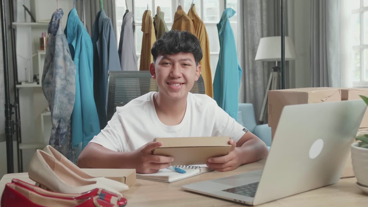 Penjual Online Pria Asia dengan Paket Melihat Kamera dan Tersenyum Sambil Menggunakan Komputer untuk Berjualan Pakaian di Rumah
