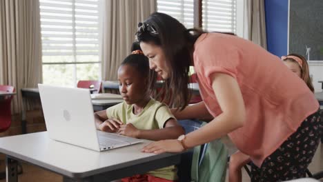 Happy-diverse-female-teacher-teaching-schoolgirls-using-laptop-in-classroom-at-elementary-school