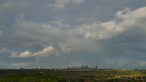 Flauschige-Wolken-Mit-Regenbogen-Nach-Regen-Mit-Skyline-In-Der-Ferne
