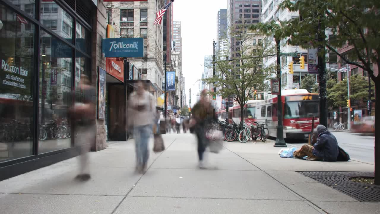 People walk briskly along the sidewalk in a bustling urban area with shops lining the street. Bicycles are parked nearby as some individuals sit on the ground, engaged in various activities.