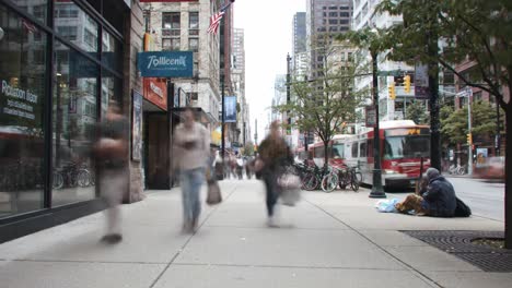people walk briskly along the sidewalk in a bustling urban area with shops lining the street. bicycles are parked nearby as some individuals sit on the ground, engaged in various activities.
