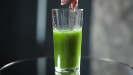 a slender hand holds a spoon in a glass full of rich, green vegetable juice