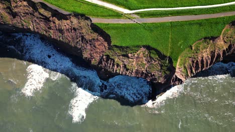 top down view of tall and large cliffside at the coast with waves crashing on the cliff wall causing white swell on a sunny day with shadows in north yorkshire seaside england