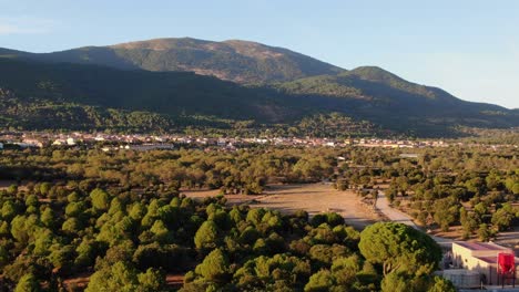 descent flight with in the golden hour of the afternoon over green pine forest with yellow summer meadows with background of forested mountains with a sunny village slopes and blue sky in avila spain