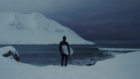 surfer standing at arctic shore with surfboard under dramatic overcast skies in iceland, hero establishing