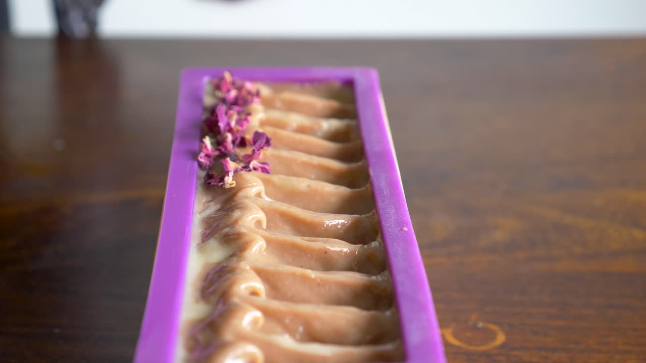 una mujer decora suavemente un bloque de jabón recién hecho con pétalos secos de una flor morada.