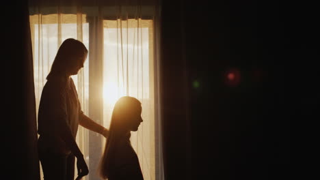 A-woman-combs-her-daughter's-long-hair.-Sitting-by-the-window-at-sunset