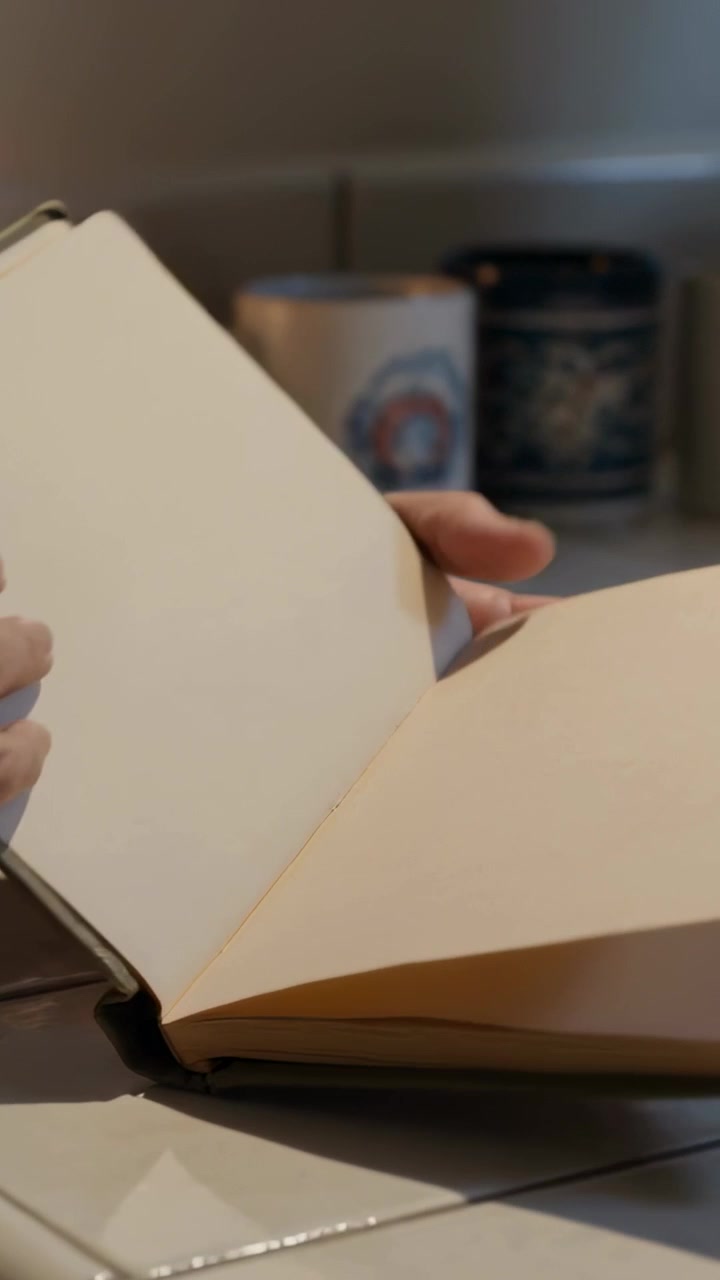 Video verticale: Mani che si inclinano con il polsino del maglione, aprono un diario in cucina vicino a una lampada, preparando appunti.