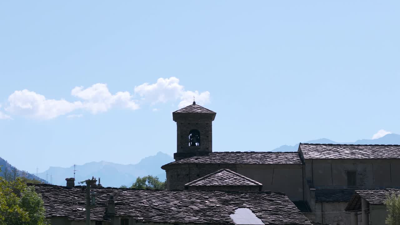 A historic bell tower rises above traditional village rooftops with majestic mountains in the background